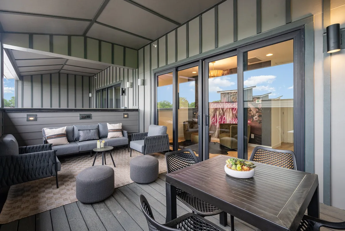 A spacious outdoor patio featuring a comfortable seating area and a dining table, with large glass doors connecting the interior to the exterior. The area is styled for relaxation or social gatherings, offering a casual and welcoming vibe that encourages group interaction. Sunset lighting through the patio adds a warm, inviting ambiance.