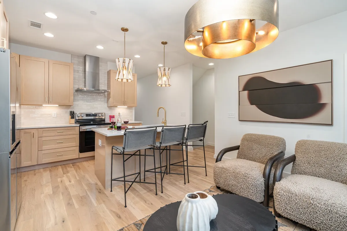 A contemporary kitchen and dining area with a spacious island and bar stools, an elegant chandelier overhead, and artwork adding visual interest. There is a designated seating area, seamlessly connecting the kitchen to a communal dining space ideal for group meals.