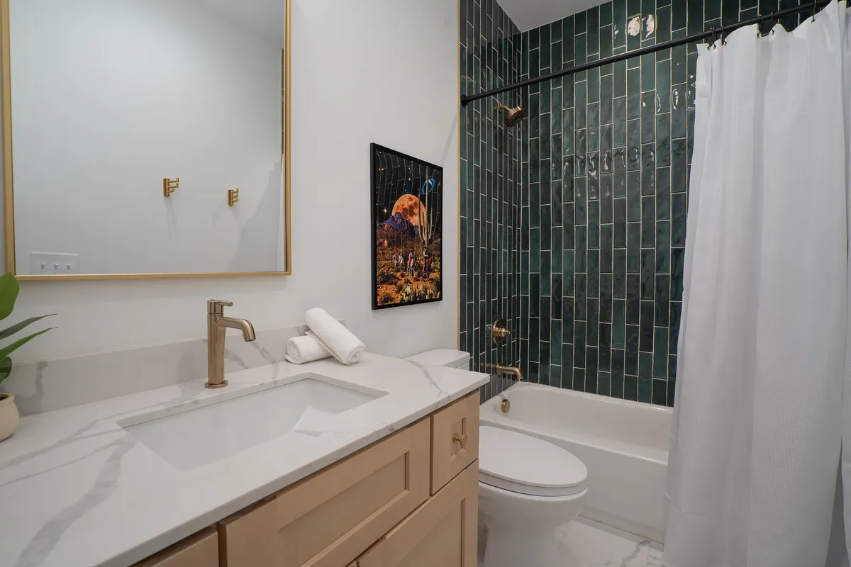 A stylish bathroom featuring a marble countertop, green tiled wall, gold fixtures, and wall-hung artwork. The environment is calm, elegant, and ideal for self-care routines.
