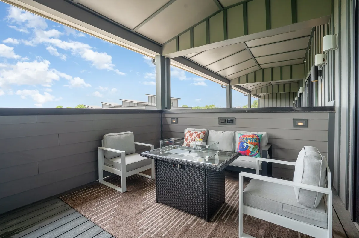 An inviting exterior patio space featuring outdoor seating with colorful decorative pillows and a glass table, suggesting a vibrant, social atmosphere. The background hints at open views, ideal for enjoying Nashville's energy.