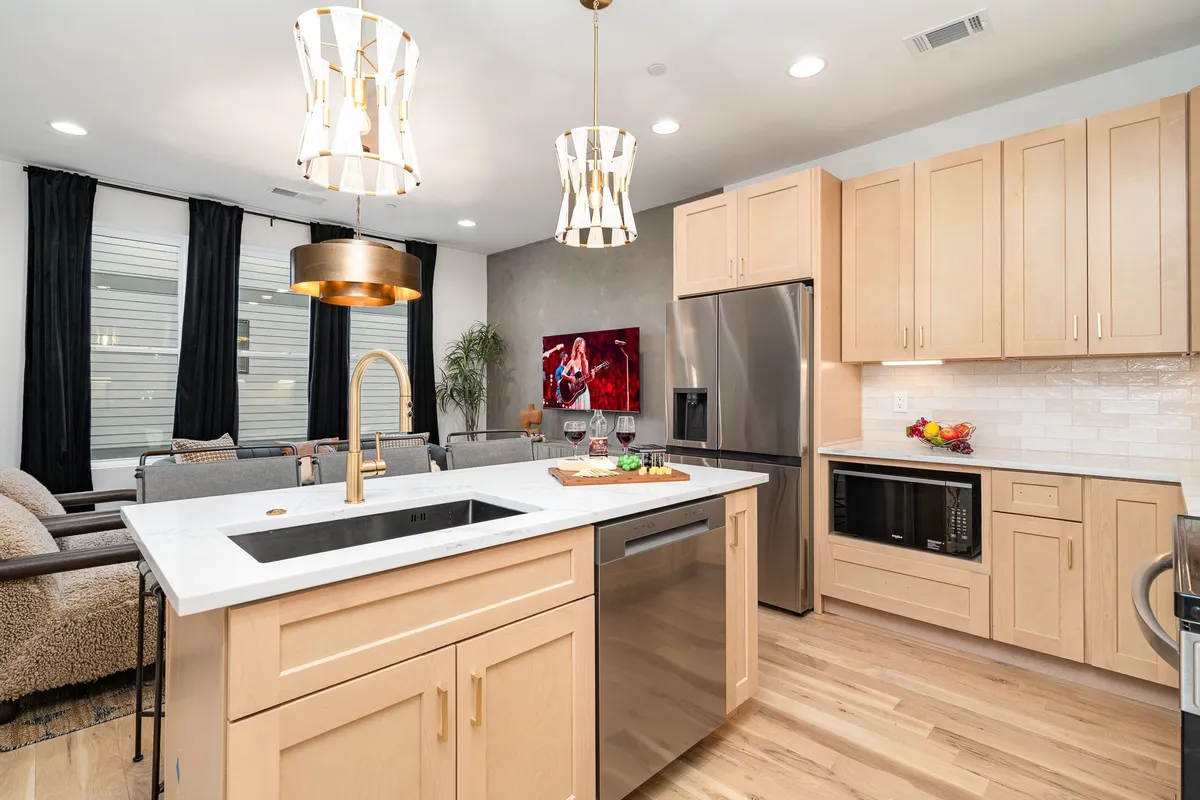 A warm, welcoming kitchen scene featuring a kitchen island with a built-in sink, stylish gold pendant lights, and stainless steel appliances. The countertop displays an appealing arrangement of food and drinks, suggesting active use and shared moments.