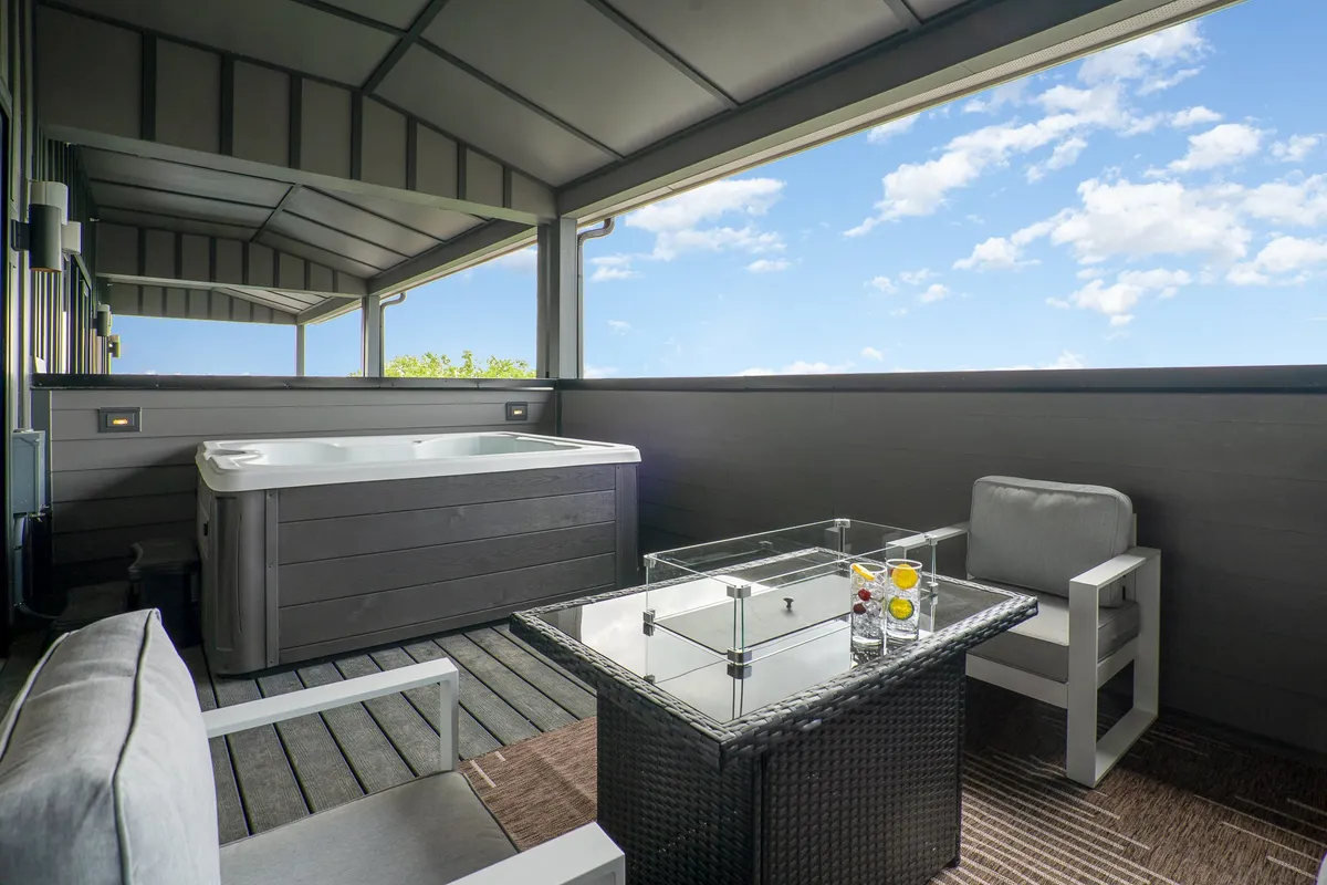 A stylish rooftop space with a hot tub, sleek glass table, and comfortable seating area, all arranged for relaxation and social interaction while enjoying sunset or city views. The setting is designed for both group festivities and private lounging.
