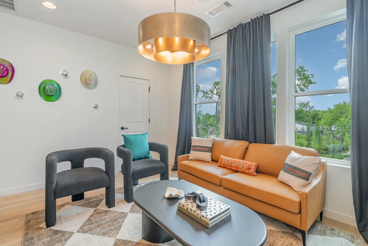 A bright and cheerful living room featuring colorful wall art, a modern light fixture, comfortable seating, and a coffee table with decor. The room is illuminated by morning light, fostering an uplifting and sociable environment for group interaction.
