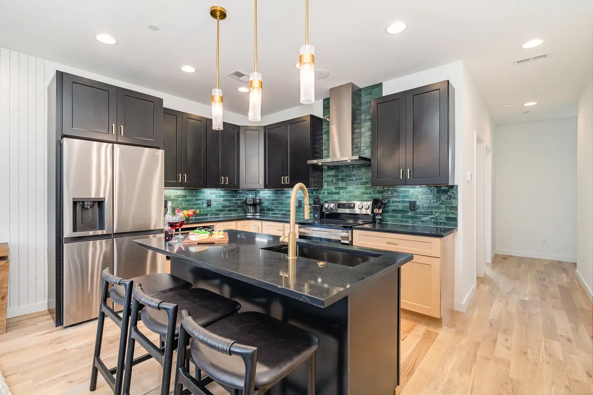 A stylish, welcoming kitchen is centered around a large island with comfortable bar stools. The space features black cabinetry, a green backsplash, and stainless steel appliances. The island is set for a social gathering, with decor and lighting that invite group cooking or mingling.