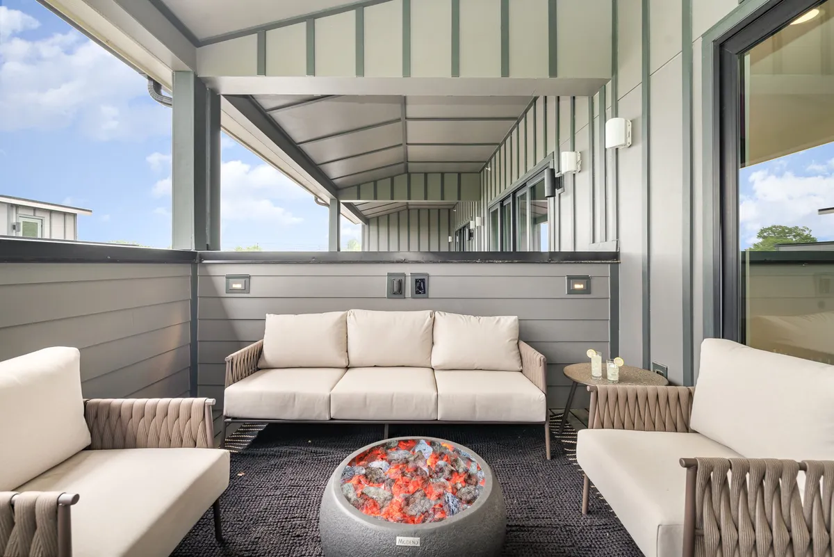 A cozy rooftop lounge area complete with a plush outdoor sofa, a glowing fire pit at its center, and stylish lighting fixtures. The open-air setting suggests a warm, inviting atmosphere ideal for intimate gatherings or unwinding beneath the stars.