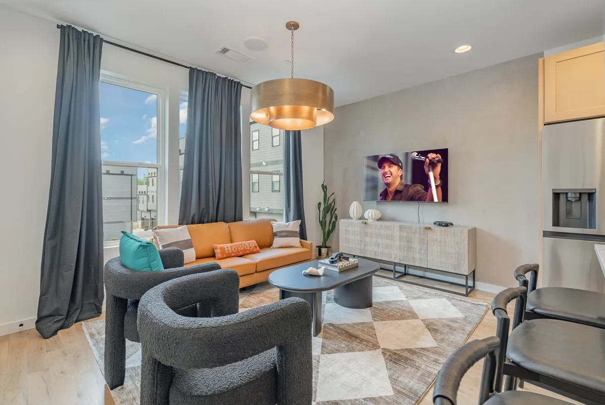 A vibrant living space with an orange sofa, blue cushion, black chairs, a TV prominently displayed, and decorative plants. The arrangement encourages both entertainment and casual conversation, supporting group activities and gatherings.