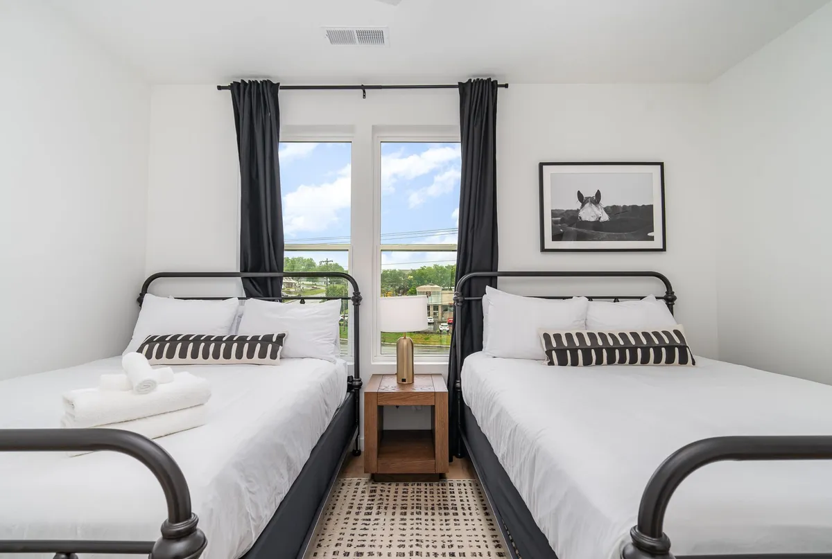 The room displays two beds with crisp white linens, large windows allowing abundant natural light, a wooden bedside table with a lamp, and serene horse artwork. The setting is calm and serene, ideal for relaxing and recharging.